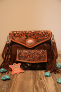 Western leather crossbody bag and wallet set with floral tooling, cowhide and fringe on rustic wood surface